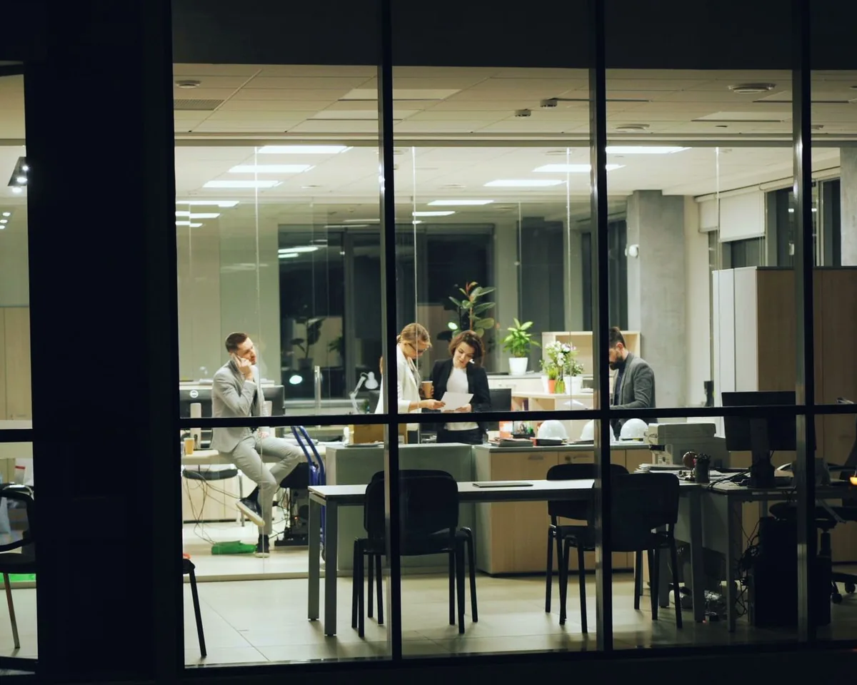 A team talking in a brightly lit office at night, seen through the building's dark windows